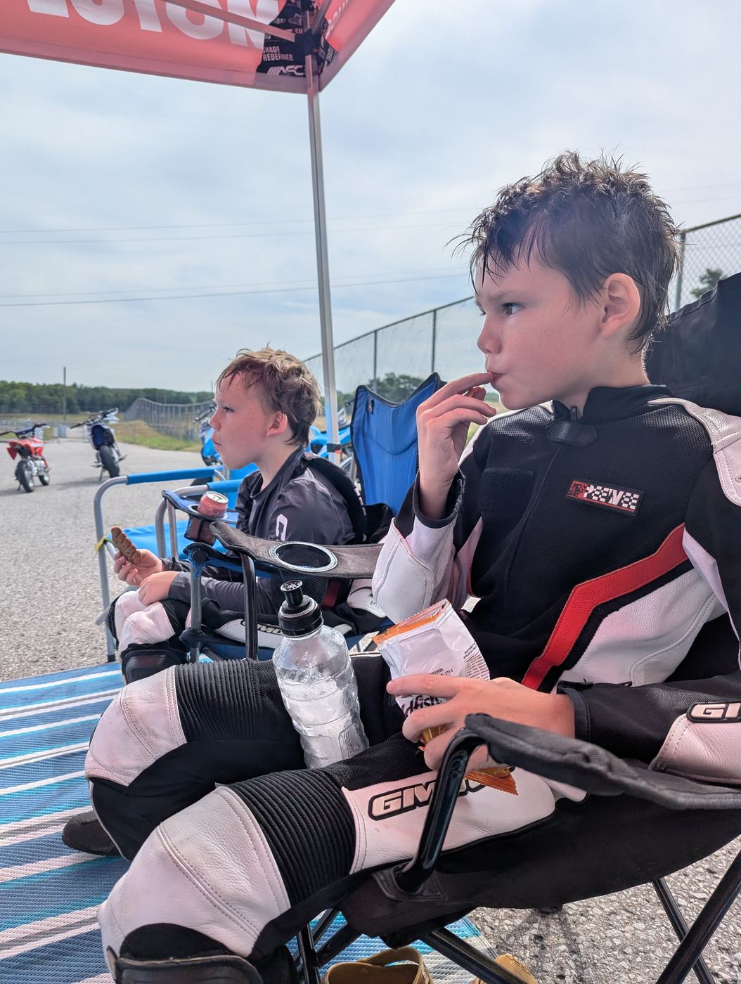 Two young riders still in their leathers, eating snacks in camp chairs under the awning between sessions