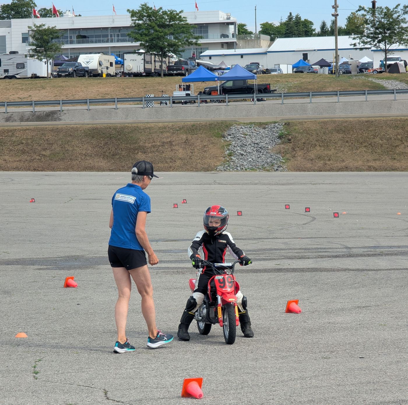 A coach in a Super Sonic shirt walking a young rider through a cone layout on the paddock tarmac at Mosport