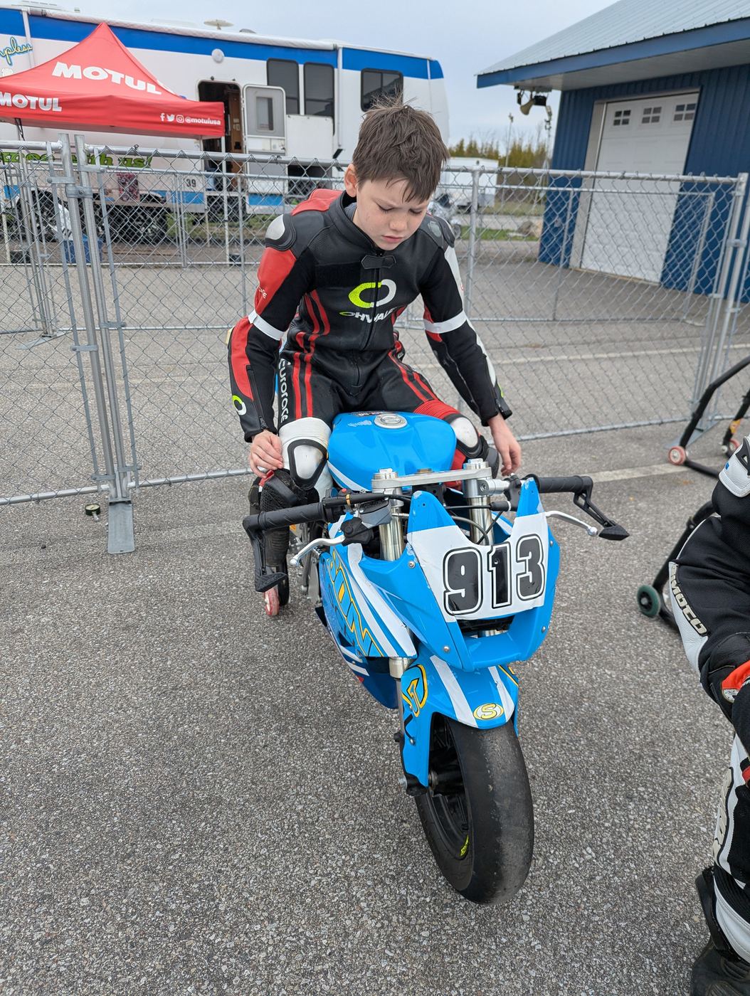 A young rider in Ohvale leathers climbing onto a blue number 913 minimoto beside the Motul awning